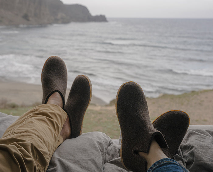 Two people relaxing with their feet up, looking out over a sandy beach. The person on the left is wearing Chestnut, brown glerups slip-on slippers, while the person on the right is wearing Chestnut, brown felted glerups boots