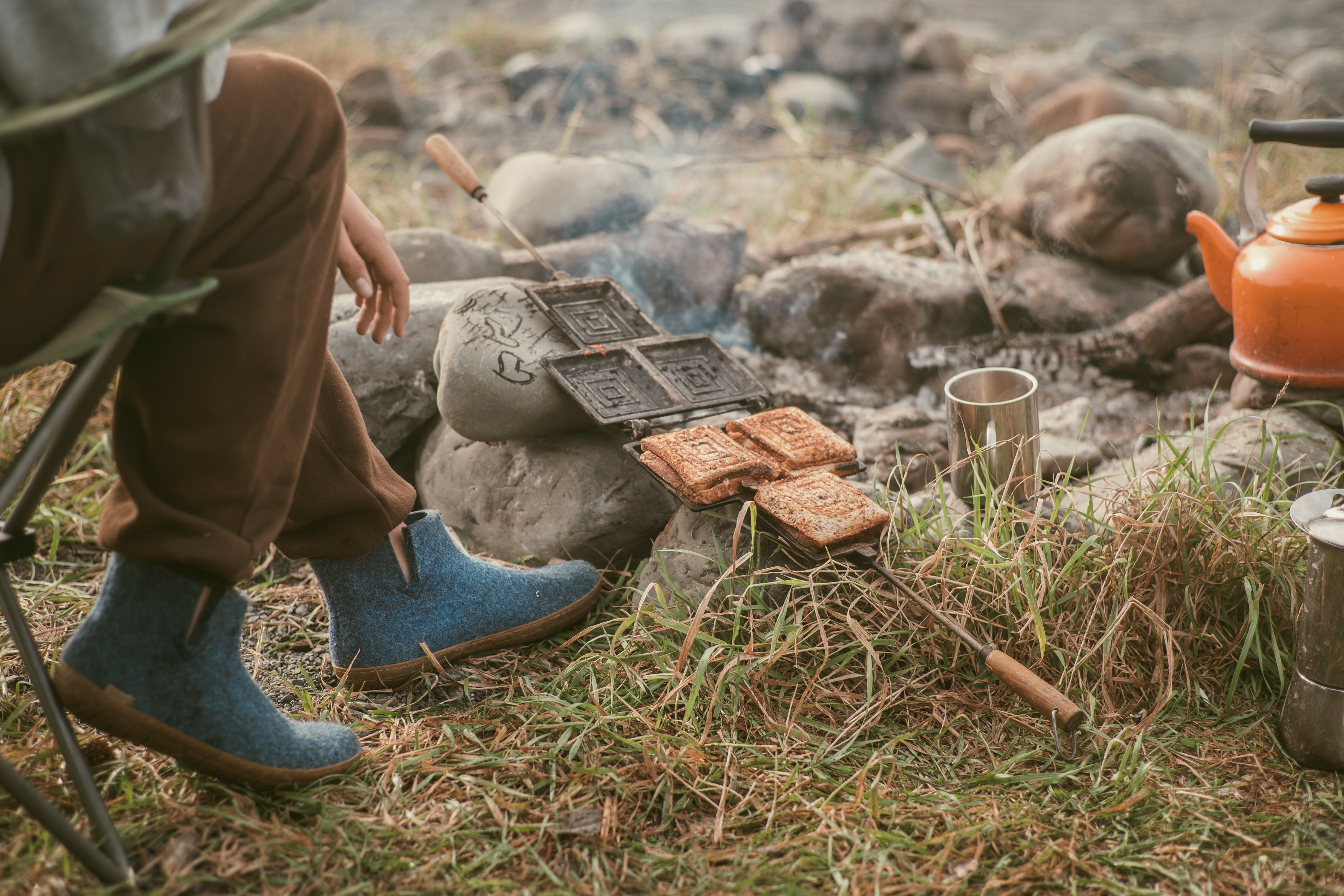 An outdoor, low-angle shot of a person sitting in a camping chair by a campfire, making toasted sandwiches in a cast-iron sandwich maker resting over the rocks, while wearing Denim, blue glerups boots with honey rubber soles