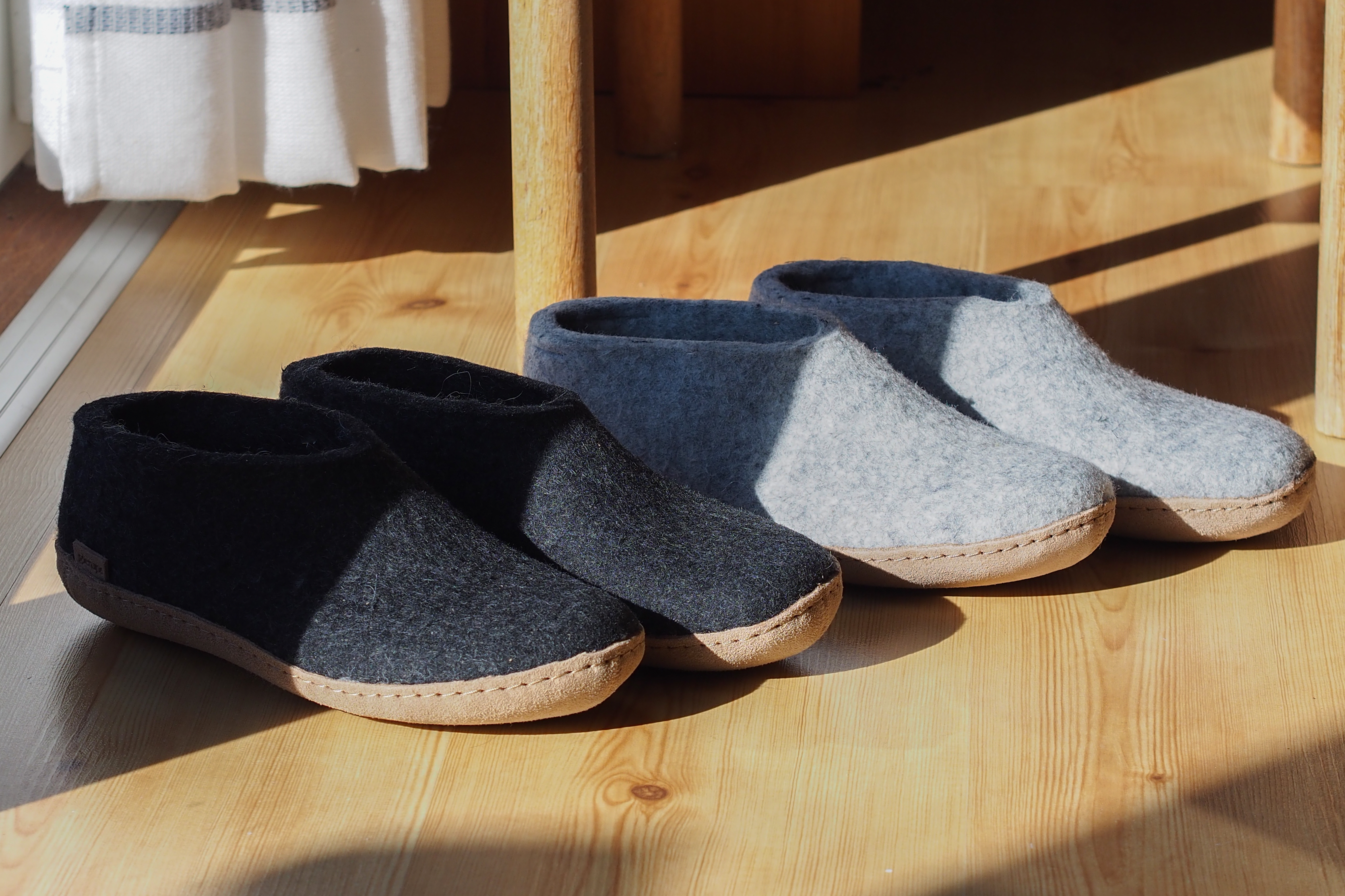 An indoor, high-angle shot of two pairs of glerups shoes sitting in a patch of bright morning sunlight on a light-colored hardwood floor near a wooden chair and a white striped curtain; one pair is charcoal, black and the other is grey, and both have leather soles.
